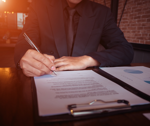 A form on a clipboard being signed by a man wearing a suit in a dark environment.