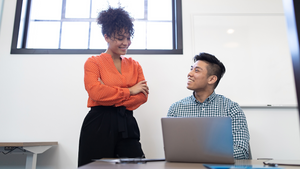 Two office workers smiling at each other while looking at a laptop.