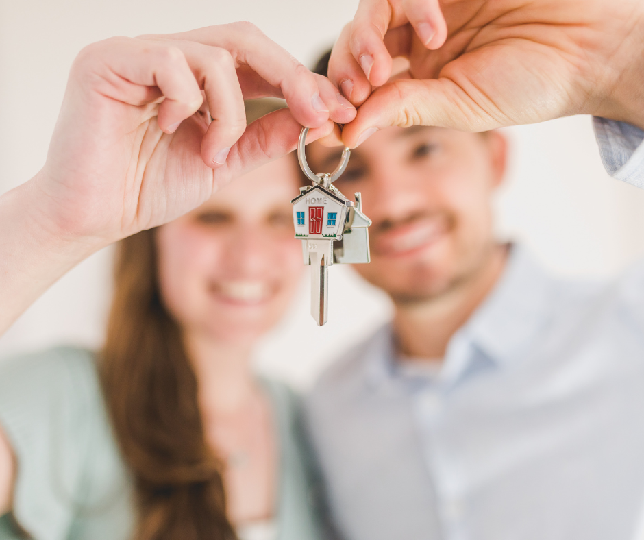 A couple holding keys together for a new house with a key ring of a house with blue windows and a red door.