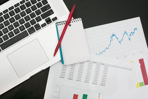 A laptop with sheets of paper in a messy pile showing graphs, charts and numbers.
