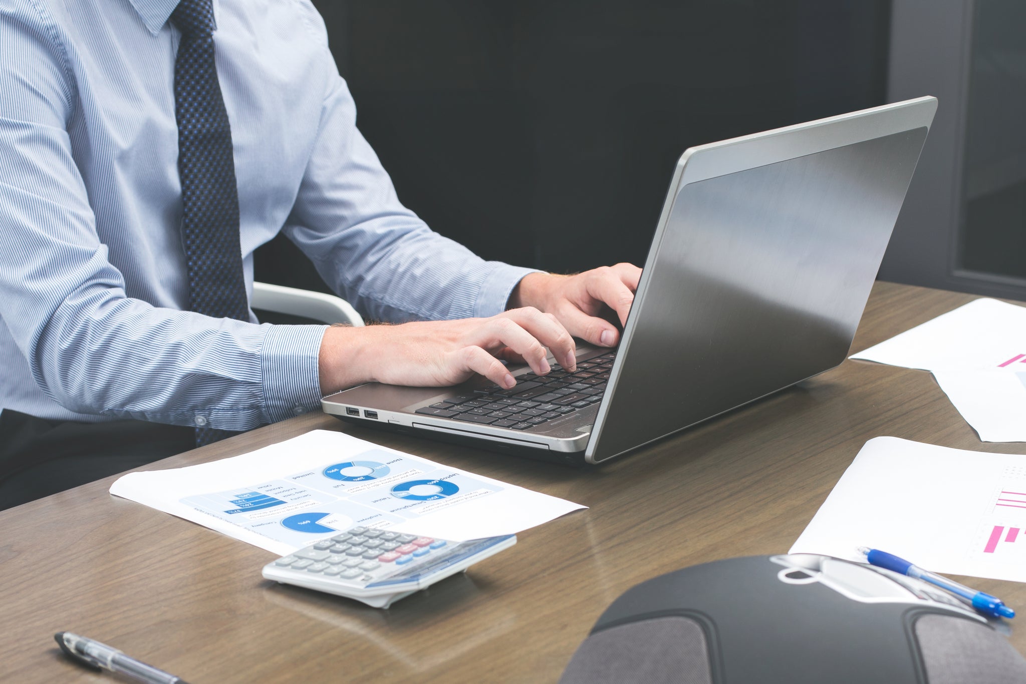 A person in a suit typing on their laptop with paperwork next to him.