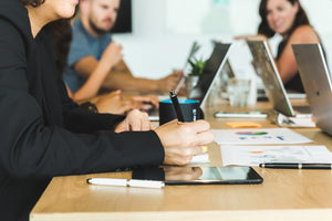 A meeting at the table with all participants having their laptops open. One person making notes with a black pen on a paper and a tablet.